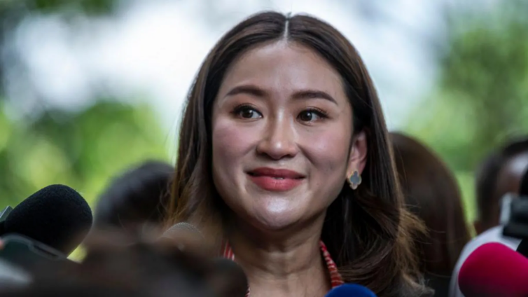 A woman with long dark hair, wearing a white shirt and earrings, smiles while standing among several microphones, suggesting she is speaking at a press conference. The background is blurred with greenery.