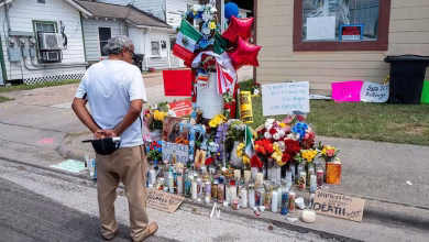 A person stands beside a street-side memorial covered with flowers, candles, signs, flags, and balloons outside residential buildings.