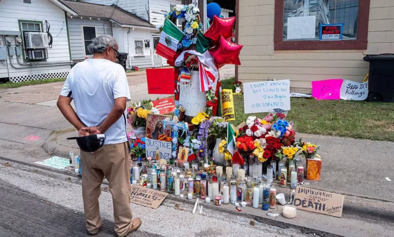 A person stands beside a street-side memorial covered with flowers, candles, signs, flags, and balloons outside residential buildings.