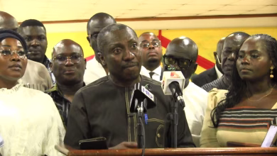 A group of people stands behind a speaker addressing microphones at an indoor press gathering.