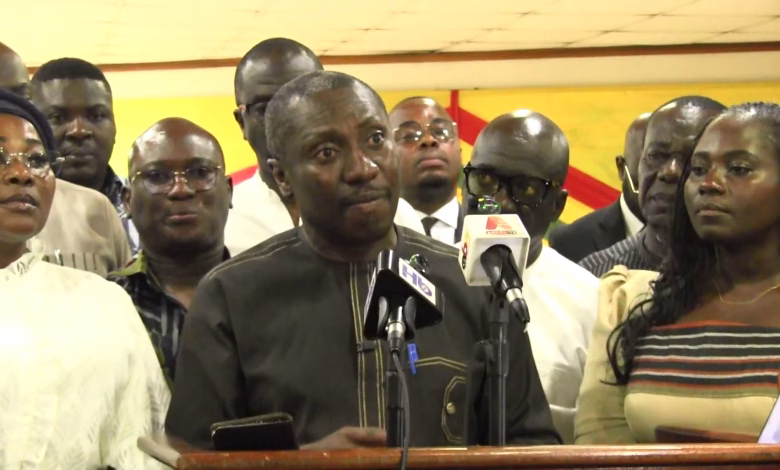 A group of people stands behind a speaker addressing microphones at an indoor press gathering.