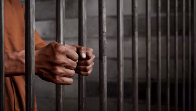A person in an orange garment grips metal prison bars inside a detention setting.