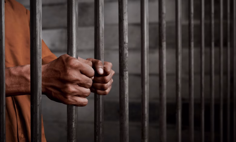 A person in an orange garment grips metal prison bars inside a detention setting.