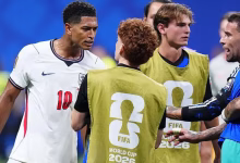 Several football players and staff stand together on the pitch, with two people wearing FIFA World Cup 2026 training bibs.