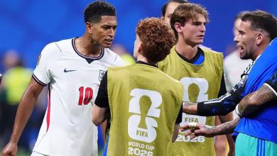 Several football players and staff stand together on the pitch, with two people wearing FIFA World Cup 2026 training bibs.