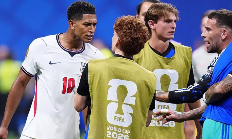 Several football players and staff stand together on the pitch, with two people wearing FIFA World Cup 2026 training bibs.
