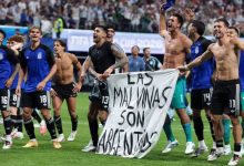 A group of football players celebrate on a pitch while holding a banner in front of a stadium crowd.