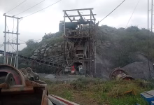 An industrial quarry site with rock-processing machinery, conveyor equipment, power lines, and piles of stone under an overcast sky.