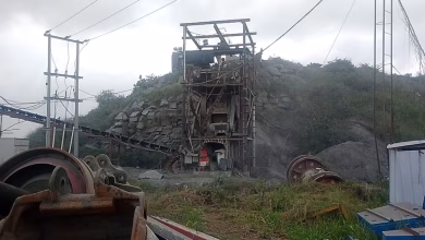 An industrial quarry site with rock-processing machinery, conveyor equipment, power lines, and piles of stone under an overcast sky.