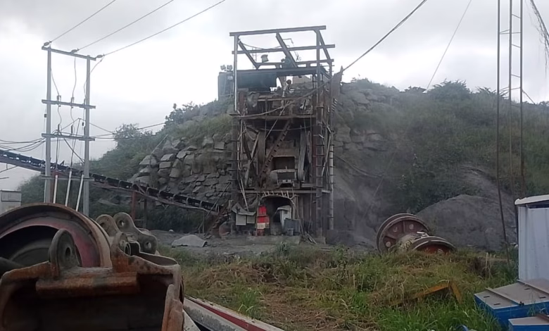 An industrial quarry site with rock-processing machinery, conveyor equipment, power lines, and piles of stone under an overcast sky.