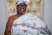 A seated older woman wearing glasses, a white headwrap, and a patterned white traditional garment is pictured against an ornate upholstered chair.