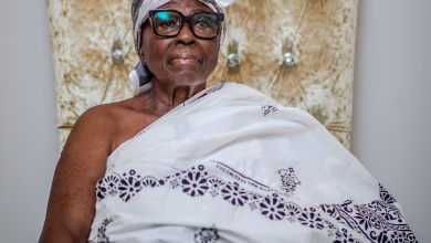 A seated older woman wearing glasses, a white headwrap, and a patterned white traditional garment is pictured against an ornate upholstered chair.