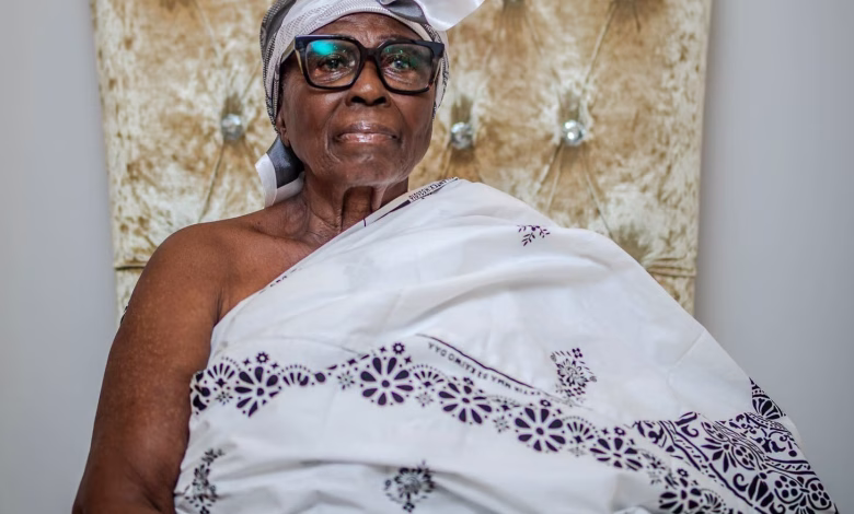 A seated older woman wearing glasses, a white headwrap, and a patterned white traditional garment is pictured against an ornate upholstered chair.