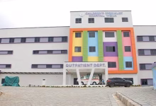 A hospital building with a brightly colored central facade, an outpatient department entrance, and parked vehicles in front.