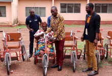 Several people stand outdoors beside a seated woman and multiple hand-powered tricycles in front of a building.