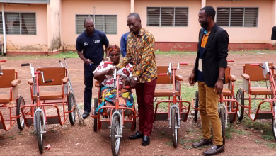 Several people stand outdoors beside a seated woman and multiple hand-powered tricycles in front of a building.
