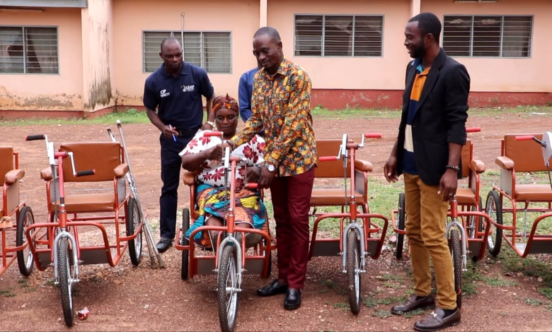 Several people stand outdoors beside a seated woman and multiple hand-powered tricycles in front of a building.