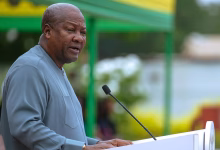 A man speaks at a podium with a microphone in front of a blurred green, yellow, and red backdrop.