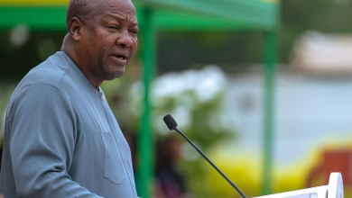 A man speaks at a podium with a microphone in front of a blurred green, yellow, and red backdrop.