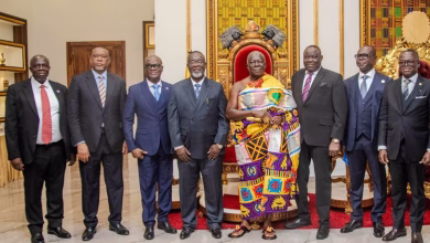 A group of men pose indoors beside ornate ceremonial chairs, with one man wearing a brightly patterned traditional cloth.