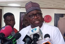A man wearing traditional attire speaks to reporters indoors while several microphones are held in front of him.