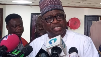 A man wearing traditional attire speaks to reporters indoors while several microphones are held in front of him.