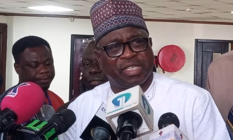 A man wearing traditional attire speaks to reporters indoors while several microphones are held in front of him.