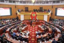 A wide view shows a parliamentary chamber with seated attendees, curved rows of desks, and a central speaker’s area.