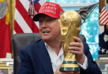 A man seated indoors holds up a gold football trophy while flags and framed items appear in the background.