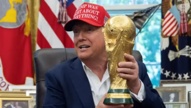 A man seated indoors holds up a gold football trophy while flags and framed items appear in the background.
