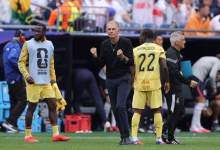 A football coach stands between two players in yellow kits on the sideline during a match.