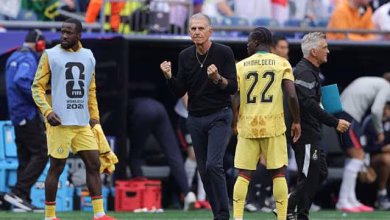 A football coach stands between two players in yellow kits on the sideline during a match.