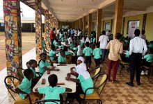 Children in green uniforms sit at tables in a covered school corridor while adults and other students stand nearby.