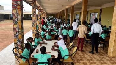 Children in green uniforms sit at tables in a covered school corridor while adults and other students stand nearby.