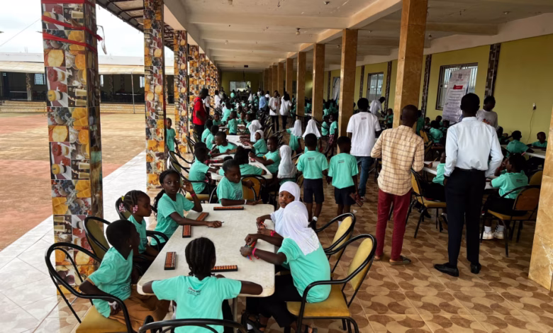 Children in green uniforms sit at tables in a covered school corridor while adults and other students stand nearby.