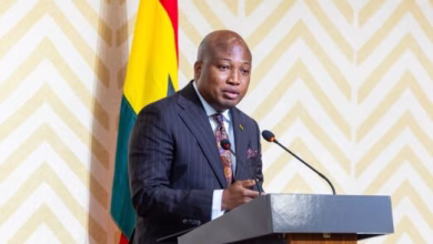 A man in a dark suit speaks at a podium with a Ghana flag behind him.