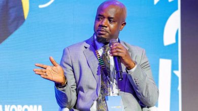 A man in a suit speaks into a handheld microphone while seated in front of a blue event backdrop.