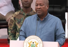 A man speaks at a podium bearing an official seal while a uniformed soldier stands behind him.