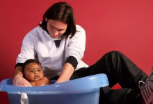 A seated adult washes a baby in a blue plastic tub against a red background.