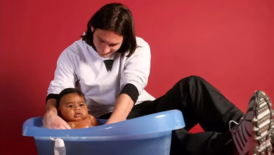 A seated adult washes a baby in a blue plastic tub against a red background.
