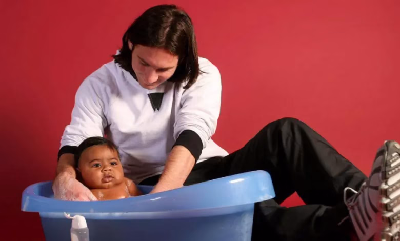 A seated adult washes a baby in a blue plastic tub against a red background.