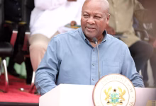 A man in a blue shirt speaks at a podium bearing a national seal, with seated people behind him.