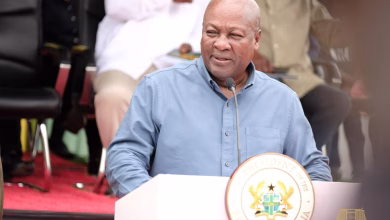 A man in a blue shirt speaks at a podium bearing a national seal, with seated people behind him.