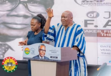 A man in a blue-and-white striped traditional outfit speaks at a podium while a woman stands beside him in front of an event backdrop.
