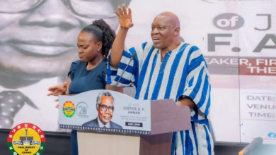 A man in a blue-and-white striped traditional outfit speaks at a podium while a woman stands beside him in front of an event backdrop.