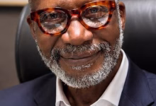 A close-up portrait shows a smiling man with glasses and a gray beard wearing a dark suit jacket and white shirt while seated indoors.