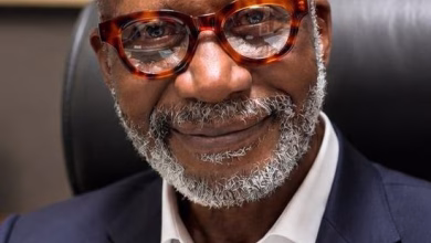 A close-up portrait shows a smiling man with glasses and a gray beard wearing a dark suit jacket and white shirt while seated indoors.