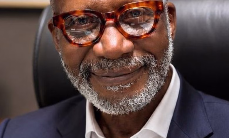 A close-up portrait shows a smiling man with glasses and a gray beard wearing a dark suit jacket and white shirt while seated indoors.