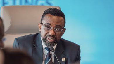 A man in a suit and glasses speaks into a microphone while seated in front of a blue backdrop.
