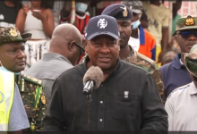 A man speaks into a microphone outdoors while surrounded by uniformed personnel and other attendees.
